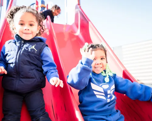 Two young students laughing as they come down a red playground slide