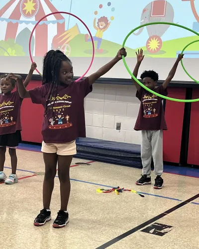 Student smiling with hula hoops raised overhead at summer camp